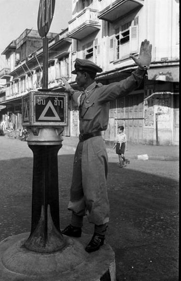 A traffic cop from back when they used to stand on elevated platforms in the middle of the intersection. The writing above the triangle says "Kap Chaa Chaa" (Drive Slowly) and on the sign at top it says "Yut" (Stop). And check out this guy's waist, what is he, a Barbie doll?